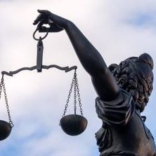 Justitia mit Wage im linken erhobenen Arm von der Seite vor blauen Himmel mit weißen Schäfchenwolken