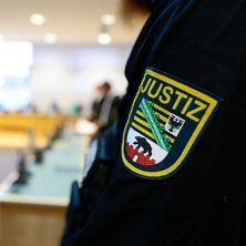 Ärmel mit Logo eines Justizbeamten im Vordergrund, dahinter verschwommen der Gerichtssaal